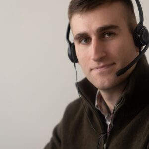 Positive young guy in casual outfit working on headphones with microphone in light workplace while looking at camera